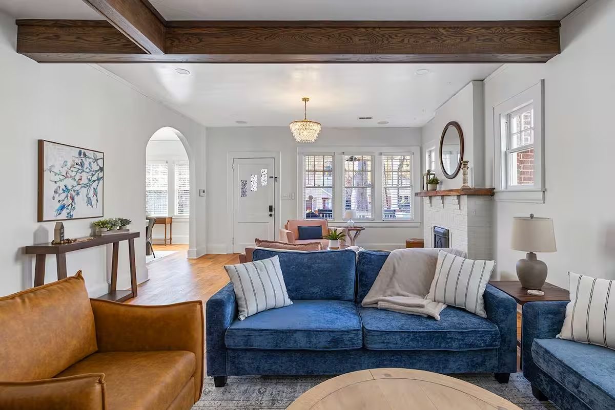 Living room with a blue velvet sofa, a cognac leather chair, exposed wood beams, and a white brick fireplace